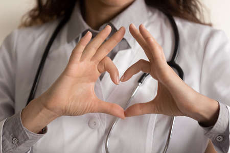 Female doctor making hand and finger heart shape, showing love sign at camera. Young cardiologist promoting medical checkup for heart disease prevention. Cardiology, healthcare concept. Close upの写真素材