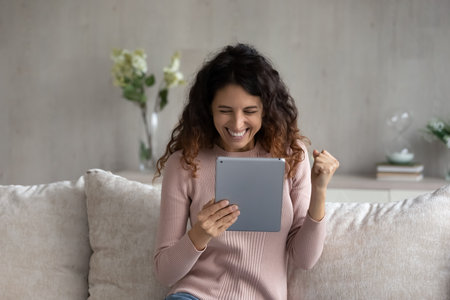 Overjoyed emotional young latina woman looking at digital tablet screen, celebrating online lottery auction betting giveaway win, feeling excited reading email with amazing news, internet success.の写真素材