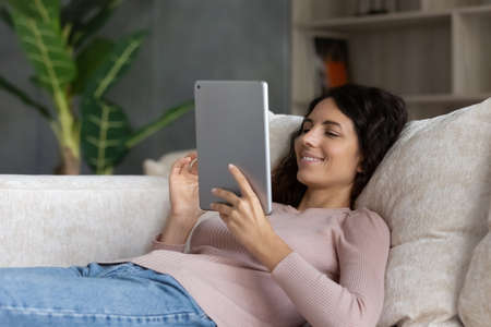 Happy lazy millennial hispanic woman using digital touchpad software applications, web surfing information, choosing goods shopping in internet store, communicating distantly lying on cozy sofa.の写真素材