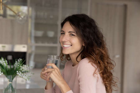 Dreamy young beautiful hispanic woman holding glass of fresh pure hot cool water sitting in kitchen,...
