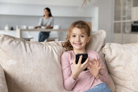 Smiling cute small child girl playing games on smartphone, communicating in social networks or watching funny videos while mother or nanny spending time in kitchen, modern technology concept.の写真素材