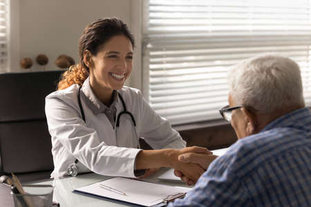 Happy female physician shaking hands with grateful old man on meeting in hospital office. Mature elder male patient thanking doctor, therapist for consultation, treatment, giving handshakeの写真素材