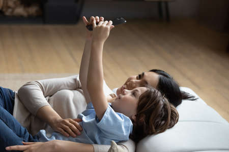 Happy little kid boy making selfie photo on web camera with caring affectionate young korean multiracial mother, recording video, streaming stories in social network, lying together on cozy sofa.の写真素材