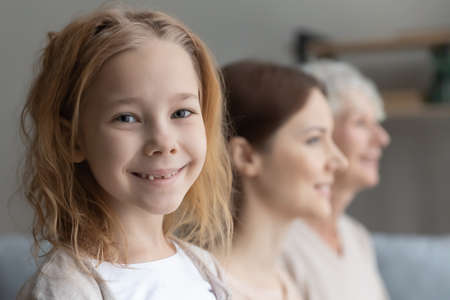 Happy cute little preschool child girl looking at camera with blurred young mother and middle aged senior grandmother, head shot close up three generations family portrait, heredity concept.の写真素材