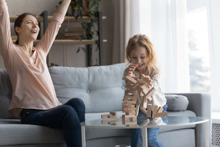 Happy emotional young mother raising arms in air, celebrating victory playing wooden board game with laughing little preschool child daughter, involved in interesting domestic activity at home.の写真素材