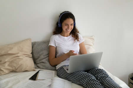 Millennial Hispanic woman sit in bed wear pyjama and headphones looks at laptop screen smile enjoy...