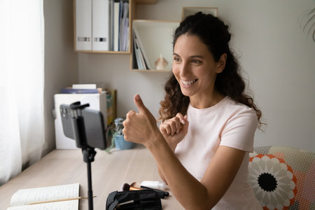 Smiling Hispanic blogger woman sit at desk record video blog on smart phone showing thumb up...