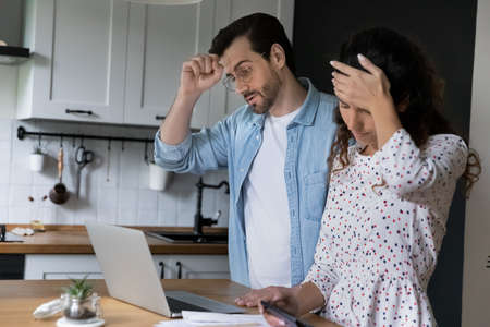 Millennial spouses standing in kitchen at home calculate household expenditures, manage family...