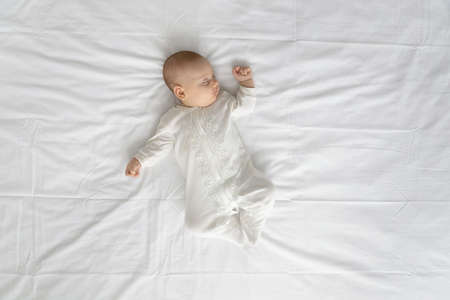 Peaceful adorable baby in bodysuit sleeping on double bed with white sheet. Top view of few month sleepy infant boy or girl lying on back mattress. Child care daily routine conceptの写真素材