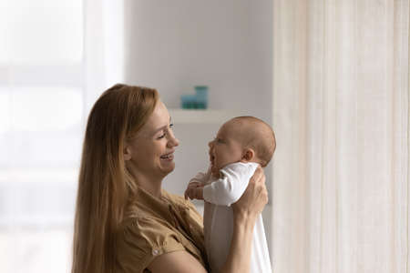 Happy mom holding adorable baby in arms, looking at little son, daughter, smiling, laughing. Young...