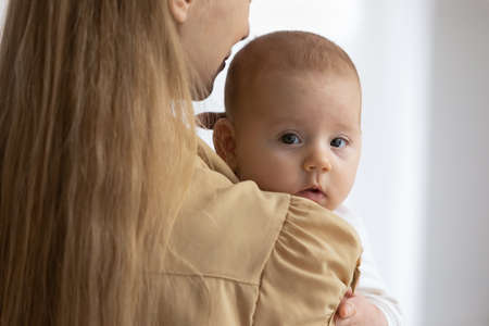 Portrait of cute little baby resting in happy mothers arms, looking at camera from behind mama...