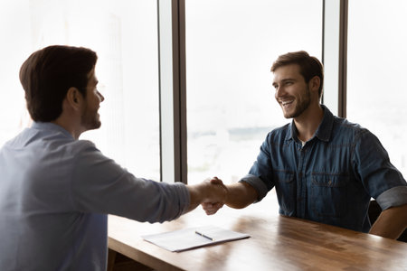 Smiling young Caucasian male employer shake hand congratulate with recruitment millennial man job candidate. Happy businessmen or business partners handshake close deal or get acquainted.の写真素材