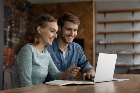 Smiling millennial man and woman colleagues sit at desk in office work together on laptop discuss business ideas. Happy diverse Caucasian coworker busy collaborate on computer. Teamwork concept.の写真素材