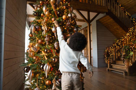 Small African American boy kid decorate big fresh Christmas fir tree with balls and garlands lights. Little teen biracial child get ready prepare for New Year winter holiday celebration alone at home.の写真素材
