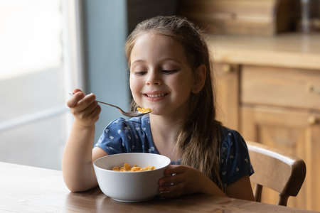 Happy small cute kid girl eating crunchy fast dry meal from bowl feeling hungry, tasting sweet corn flakes with milk on breakfast, sitting at kitchen table, balanced meal for children concept.の写真素材