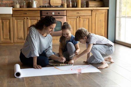 Happy millennial mother or nanny teaching little siblings kids drawing with brushes on poster paper, enjoying creative art family activity, sitting on heated warm wooden floor together in kitchen.の写真素材