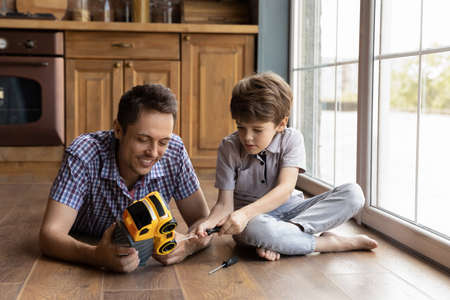 Smiling young father teaching little son repairing toy, playing together on warm floor in kitchen. Adorable small concentrated kid boy fixing car using screwdriver, improving skills with dad at home.の写真素材