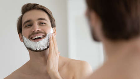 Young smiling Caucasian shirtless man applying shaving foam on face in front of mirror, preparing for hair removal morning routine. Concept of personal hygiene, skincare, male facial beauty productsの写真素材