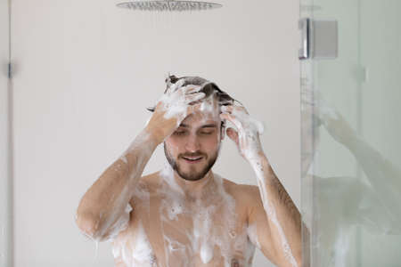 Calm handsome 30s man with foamy head and body taking a shower, wash hair with anti dandruff shampoo...