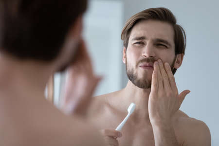 Handsome young man look in mirror touch cheek with hand, feels pain while brushing teeth due cavity or caries. Toothache, dental check up, oral care, need toothpaste for sensitive tooth enamel conceptの写真素材