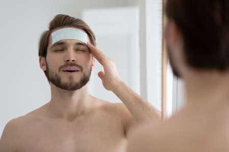 Young shirtless man apply facial anti-wrinkle, moisturizing patches, smoothing cleansing line pad on forehead, caring for face skin, enjoy procedure, improves complexion, hygiene and skincare conceptの写真素材