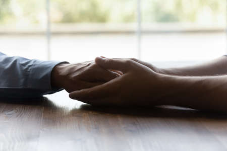 Crop close up grownup son holding senior father hands, expressing love and care, sitting at wooden...