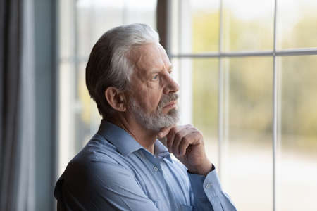 Head shot thoughtful upset mature 60s man looking out window at home alone, touching chin, lost in...