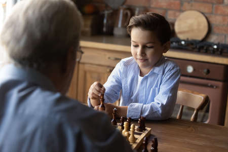 Little grandson with senior grandfather playing chess at home together close up, family enjoying...