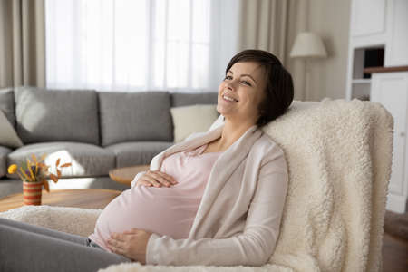 Sweet moments of anticipation. Charming expectant mother relax in comfy chair at living room dream about happy future life imagine newborn baby. Smiling young pregnant woman rest at home touch tummyの写真素材