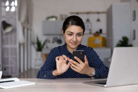 Confident Indian businesswoman using smartphone, sitting at home office desk with laptop, young woman freelancer or student typing and looking at phone screen, distracted from work, chatting onlineの写真素材