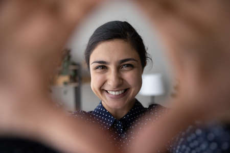 Head shot portrait smiling Indian woman having fun with camera, looking through fingers gesture,...