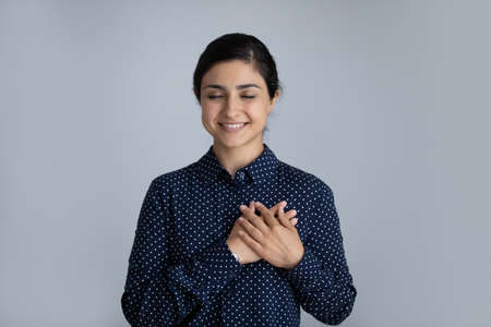 Head shot smiling Indian woman with closed eyes holding hands on chest, standing on grey studio background isolated, happy young female thanking god and faith, feeling love gratitude or making wishの写真素材