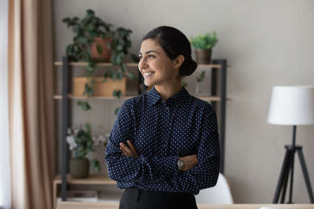 Profile smiling dreamy Indian businesswoman looking in distance, standing with arms crossed, happy young female, student or freelancer visualizing good future, dreaming of new job opportunitiesの写真素材