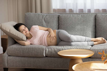 Cute moments of pregnancy. Happy serene future mother lying on sofa in comfy posture holding palm on naked big belly enjoy quiet moment of peace. Carefree pregnant woman sleep dream nap at living roomの写真素材