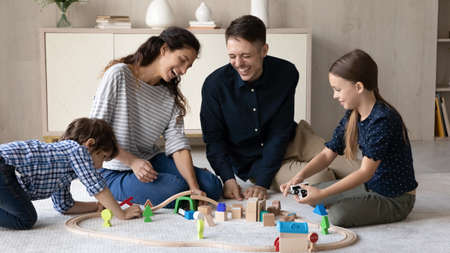Happy young hispanic couple parents having fun with little adorable kids son daughter playing wooden toys railway road, sitting together on floor carpet, joyful family entertaining in living room.の写真素材