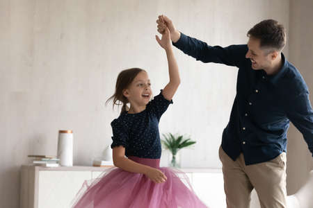 Happy young latin father twisting laughing cute little child daughter dancing to funky music together in modern living room. Smiling two generations family having fun, enjoying carefree weekend time.の写真素材