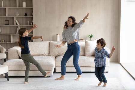 Happy beautiful young hispanic mother dancing barefoot with joyful adorable little children siblings in modern living room, homeowners enjoying carefree weekend domestic activity together at home.の写真素材