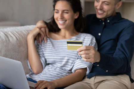 Focus on credit card in hands, happy young married family couple buyers entering payment information purchasing goods or services in internet store on computer sitting on sofa, online shopping conceptの写真素材