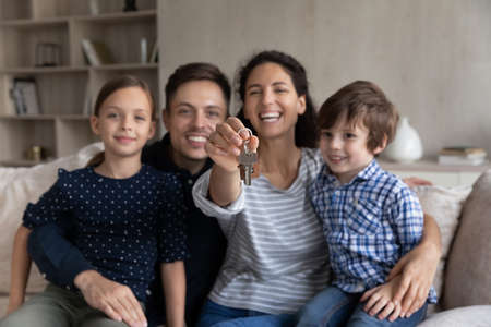 Portrait of smiling young hispanic family couple and cute little children showing keys, sitting on cozy sofa, feeling excited of moving into new own apartment, celebrating purchasing new dwelling.の写真素材