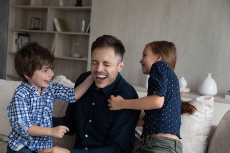 Joyful small children boy girl tickling laughing young latin daddy, having fun together on cozy sofa in living room, happy two generations family playing entertaining together on weekend at home.の写真素材