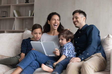 Joyful happy two generations full family laughing at funny story, reading book at home. Happy young hispanic couple parents involved in domestic hobby activity with small children son daughter.の写真素材