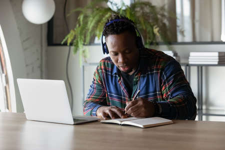 Focused serious African American student guy in headphones watching, listening to webinar on laptop,...