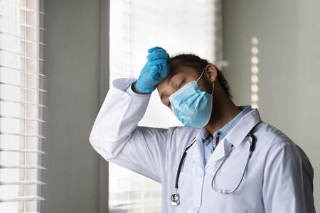 Exhausted young doctor feeling fatigue, stress due to work in hospital. Tired African mixed race practitioner in medical face mask, protective gloves wiping sweat from forehead with closed eyes.の写真素材