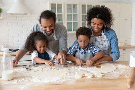 Happy African American parents with kids cooking homemade cookies in kitchen together, using cutters, loving young mother and father with little son and daughter spending leisure time at homeの写真素材