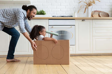 Happy loving African American father and daughter playing together, family engaged in funny activity, caring young dad pushing cardboard box with 5s girl child holding toy spyglass, having funの写真素材
