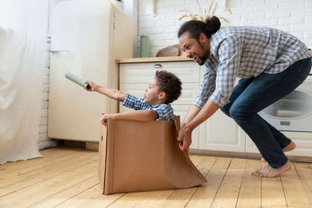 Excited laughing African American father pushing cardboard box with little son, happy family playing funny game, loving smiling young dad and 5s boy child having fun in kitchen at home togetherの写真素材