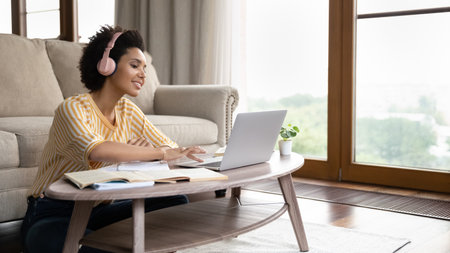 Smiling young african american multiracial woman in headphones watching educational lecture on computer, preparing for exams, sitting on floor at coffee table at home, copy space for e-learning ad.の写真素材