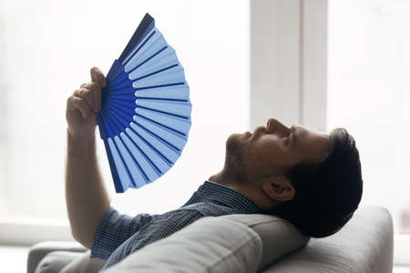 Profile view tired overheated man waving paper fan, cooling himself, suffering from heat in room without conditioner, exhausted young male leaning back resting on couch at home, breathing fresh airの写真素材