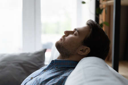 Close up profile of calm man resting leaning back on cozy couch at home, daydreaming or taking day nap, relaxed tired young male with closed eyes spending lazy leisure time, sleeping on sofaの写真素材