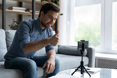 Smiling young man showing thumb up, using smartphone on tripod, satisfied blogger influencer...